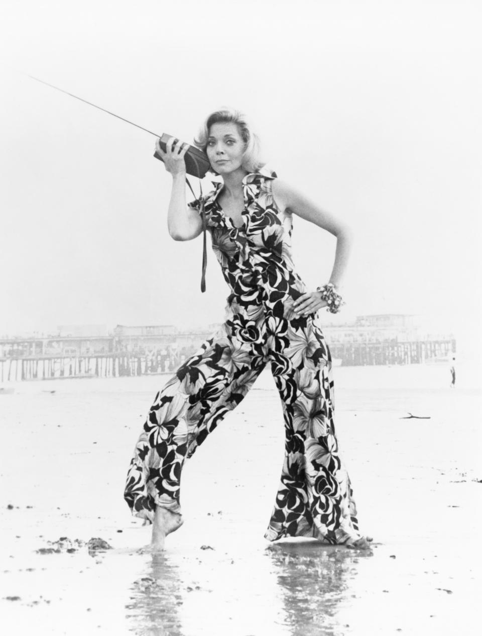 Elaine Stewart poses on a beach holding a vintage portable radio to her ear, wearing a floral-patterned jumpsuit. A pier is visible in the background