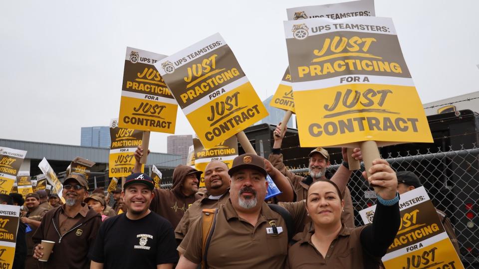 UPS workers in brown uniforms standing outside a warehouse, holding signs that say 