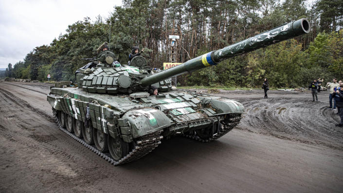 Ukrainian soldiers ride a tank down an empty road as photographers take pictures of it.