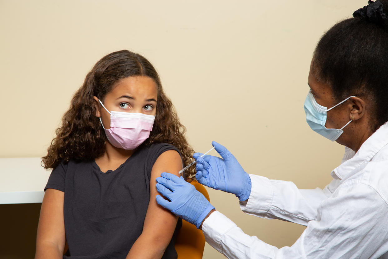 A young person receives a shot to the upper arm, given by a gloved health care worker while both wear face masks.