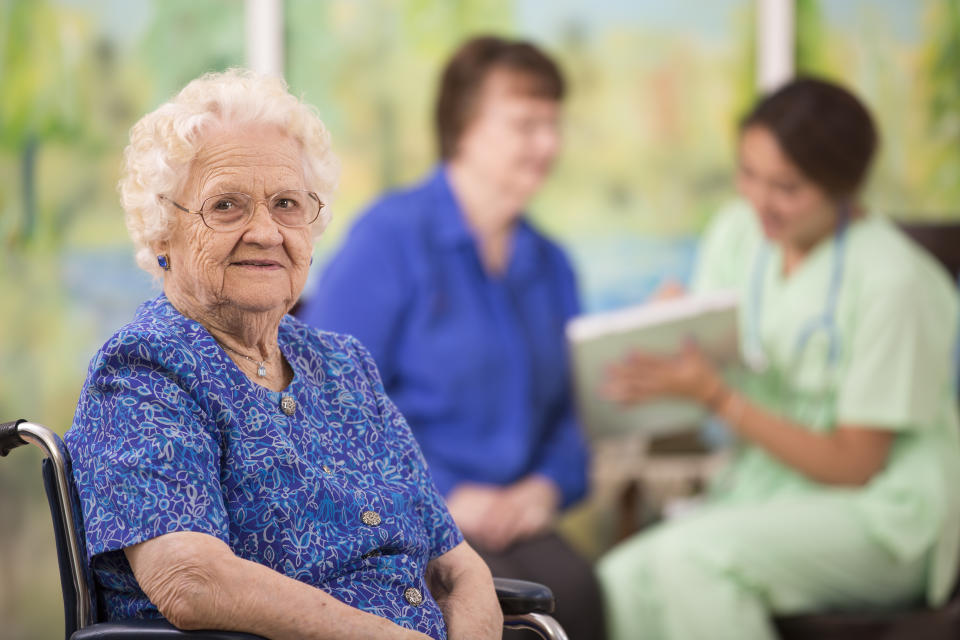 Paciente femenina de 100 años en primer plano con su enfermera de atención domiciliaria de ascendencia latina en segundo plano, discutiendo opciones de tratamiento con la hija en una residencia de ancianos o en el entorno del hogar.