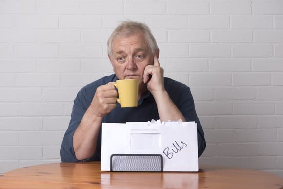 A visibly worried senior man drinking coffee in front of a stack of bills.