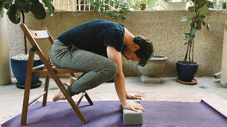 Man on a chair practicing Crow Pose by leaning forward with hands on blocks