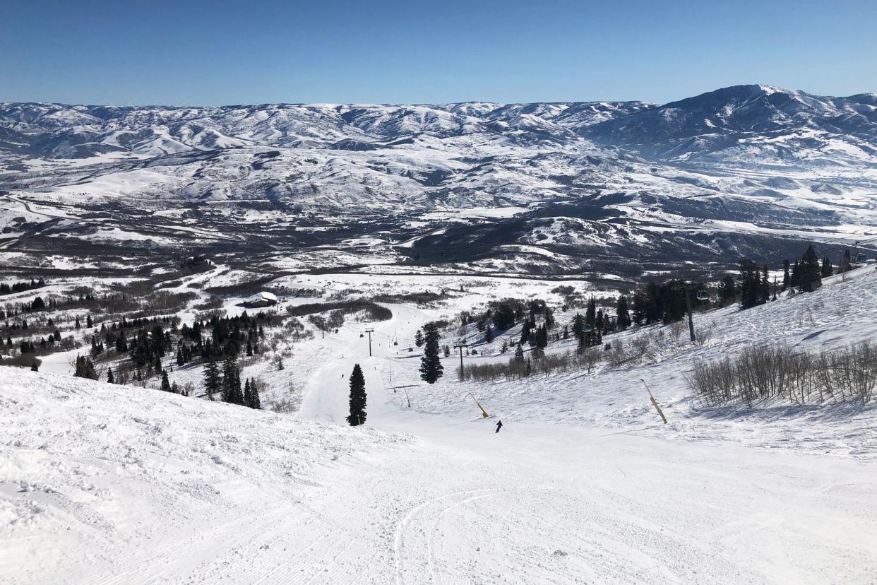 Looking down a slope at Snowbasin Ski Resort, Ogden, Utah, surrounded by snow-covered mountains, bright snow, mountains in the distance with a light blue sky