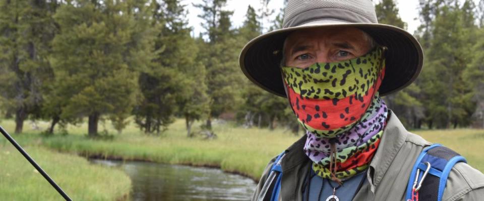 Attractive older Caucasian man with blue eyes, with face covered by bandana. Standing beside a river with fishing rod, trees and grass in background. He looks like a retiree.