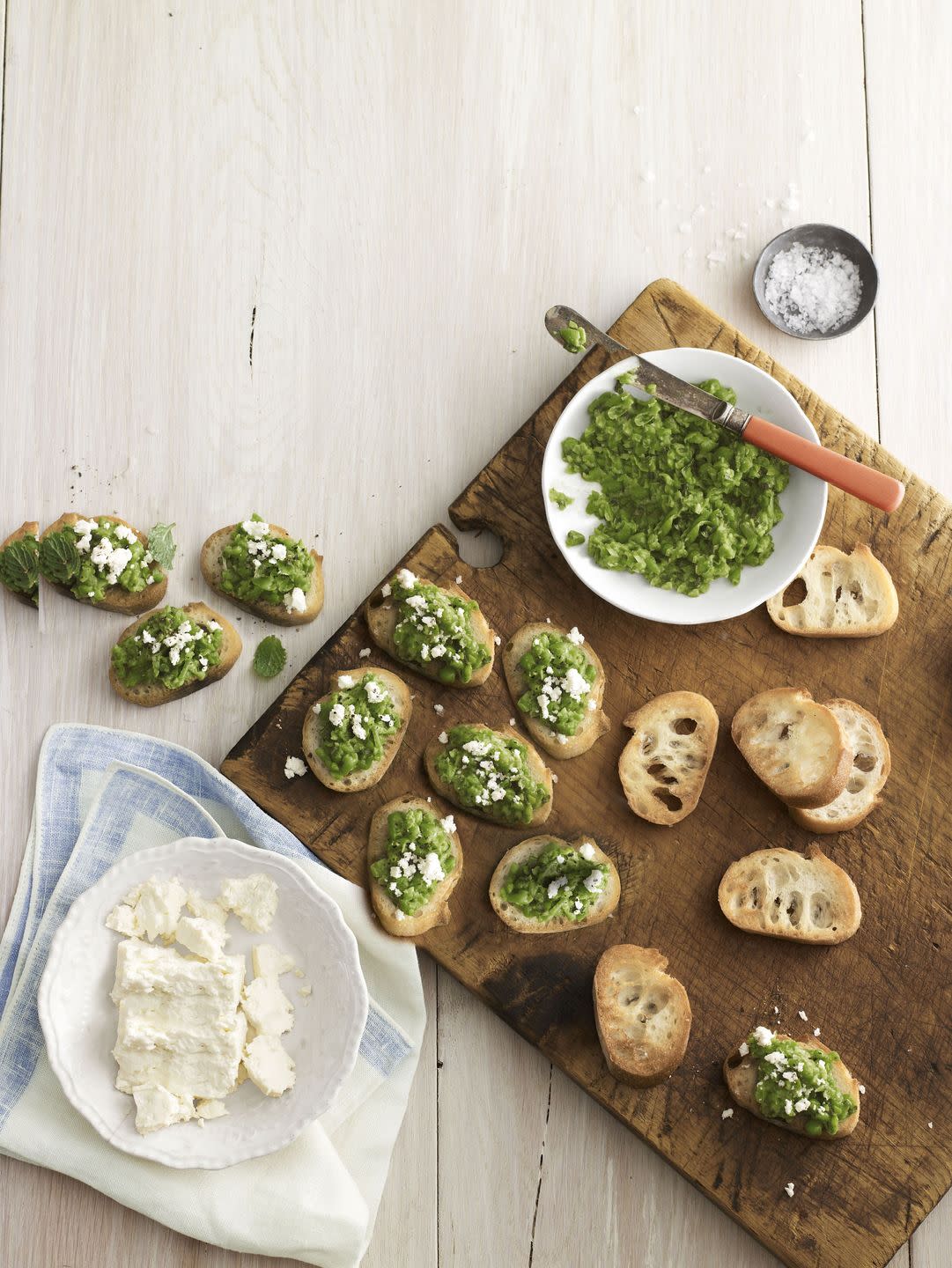 pea and feta crostini arranged on a wooden serving board with small bowls of pea spread and feta cheese
