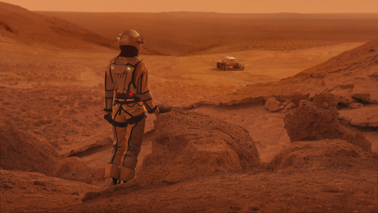  Red dirt on Mars surface. Woman wearing futuristic exoskeleton exploring cave and mountain areas. 