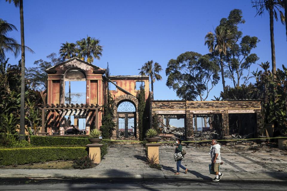 Two people walk by a home whose front is still standing but the rest of which is burned down