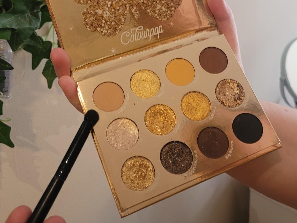 The writer holds the Colourpop Good as Gold eye-shadow palette and holds a black brush