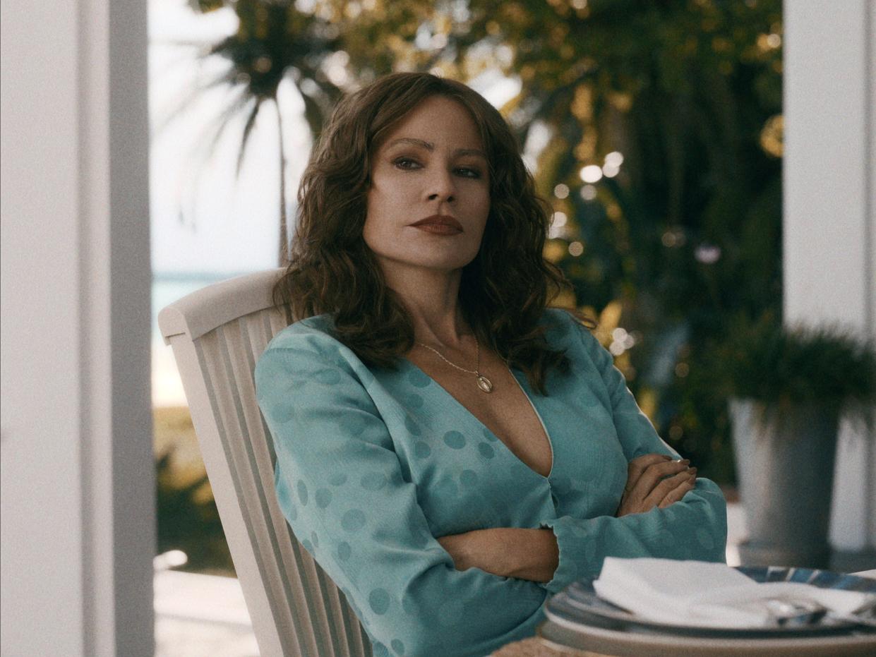 sofia vergara as griselda in the netflix series. she's wearing a light blue, low cut blouse, sitting at an outdoor table in a tropical setting with a stern expression on her face and her arms crossed
