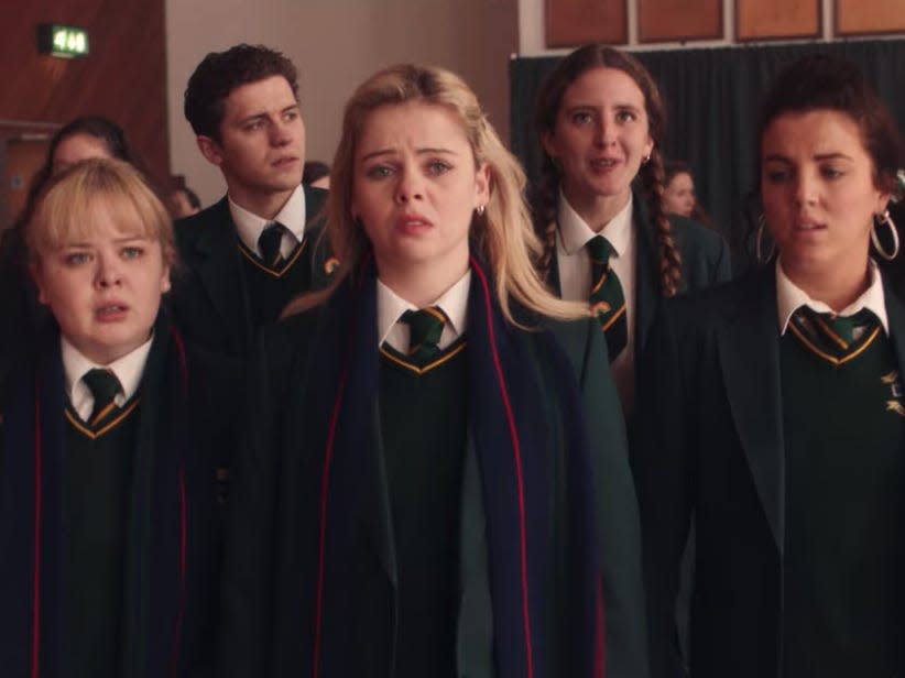 a group of five students walking in a school gymnasium, dressed in green and yellow school uniforms in derry girls