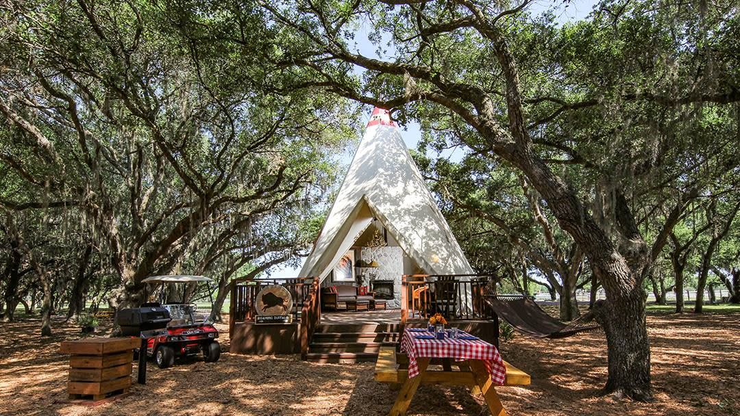 a teepee sits atop a front porch in a field of oak trees with a picnic table nearby