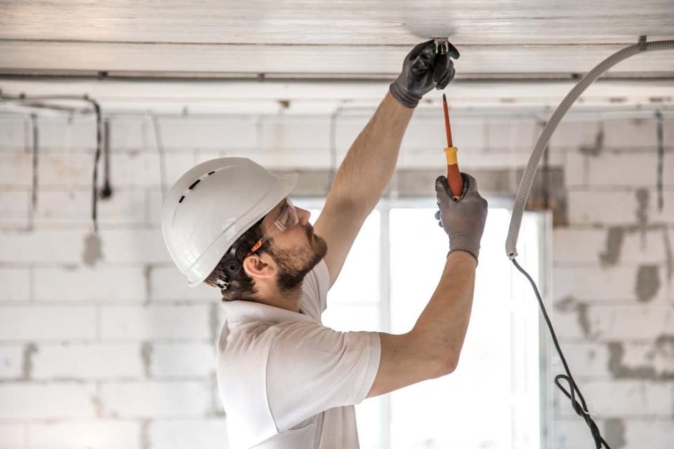 A man in a white shirt and a white hard hat used a tool to fix wires on a ceiling. 