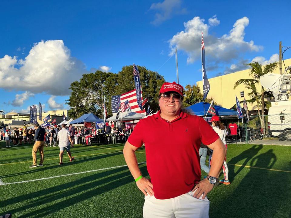 A man in a MAGA hat standing in a grassy field, hands on his hips, with people and merchandise tents behind him