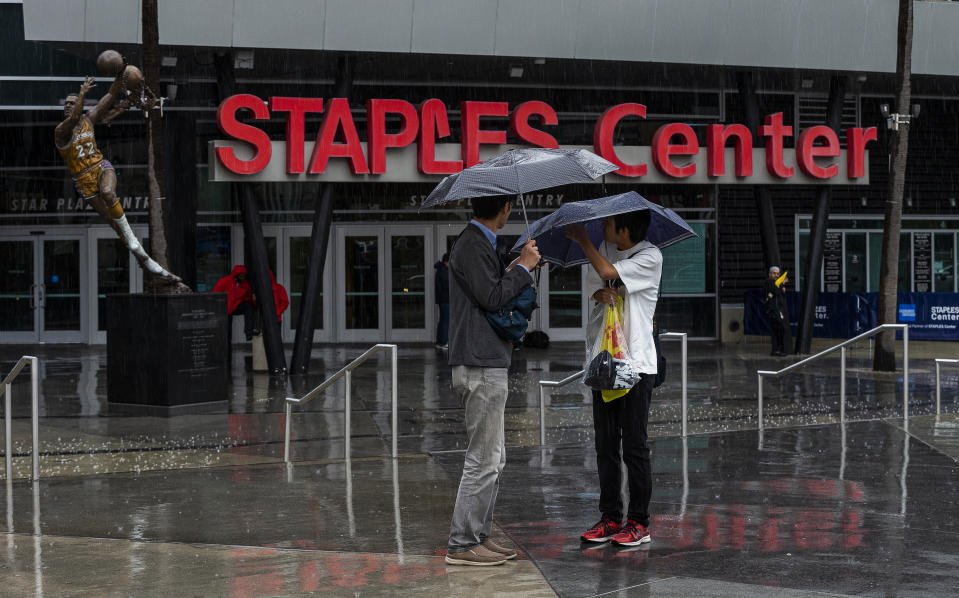Tourist stand under umbrellas in rain outside Staples Center, home to hockey's Los Angeles Kings and basketball's Los Angeles Lakers, Clippers and Sparks, in Los Angeles on Thursday, March 12, 2020. Officials have banned large gatherings and events in Los Angeles to try to stop the spread of the new coronavirus. (AP Photo/Damian Dovarganes)