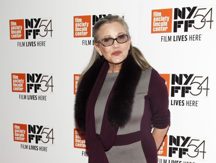 Carrie Fisher wearing tinted glasses, a purple and grey dress and a furry scarf
