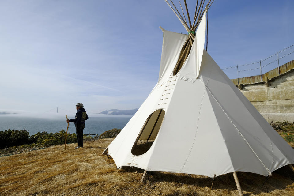 In this photo taken Tuesday, Nov. 12, 2019, Eloy Martinez, who took part in the Native American occupation of Alcatraz 50 years earlier, stands by a teepee on the island and looks out toward the bay and Golden Gate Bridge in San Francisco. The week of Nov. 18, 2019, marks 50 years since the beginning of a months-long Native American occupation at Alcatraz Island in the San Francisco Bay. (AP Photo/Eric Risberg)
