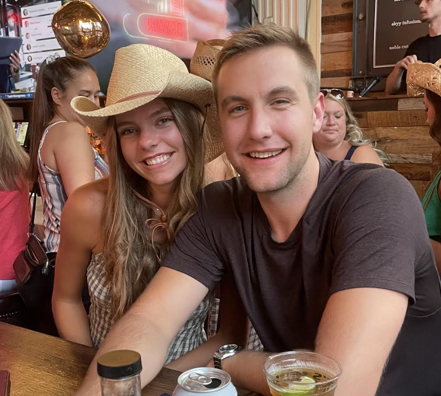 Two individuals smiling at a bar, man with beer and woman wearing a straw hat, with a sports game on TV in the background