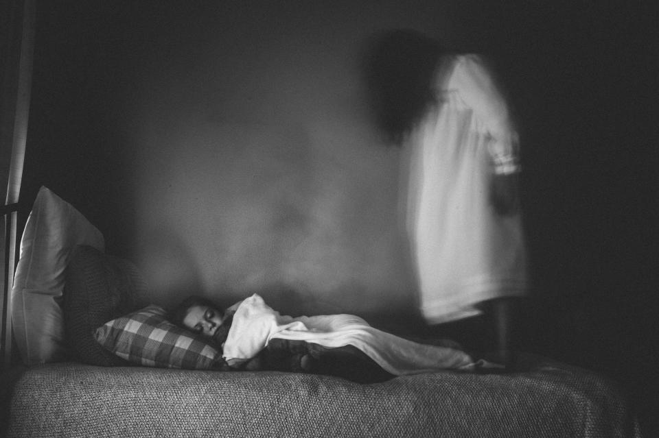 A girl sleeping in bed with a ghostly figure of a woman in a nightgown and dark hair covering her face, standing on the end of the bed above the girl's feet