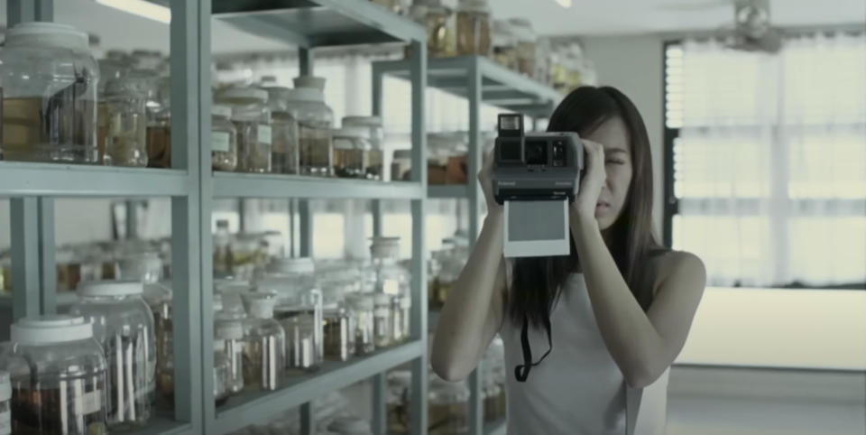 A woman is taking a picture with a vintage instant camera in a laboratory filled with jars on metal shelves