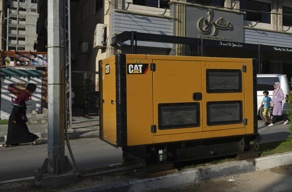 People walk near an electricity generator used by a restaurant, on the main road in Gaza City, July 8, 2022. Private generators are ubiquitous in parts of the Middle East, spewing hazardous fumes into homes and business across the country, almost 24 hours a day. As the world looks for renewable energy to tackle climate change, Lebanon, Iraq, Gaza and elsewhere rely on diesel-powered private generators just to keep the lights on. The reason is state failure: In multiple countries, governments can’t maintain a functioning central power network, whether because of war, conflict or mismanagement and corruption. (AP Photo/Adel Hana)