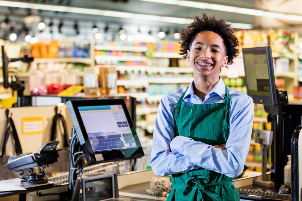 A teenage cashier smiles at the camera while standing behind the cashier counter. 