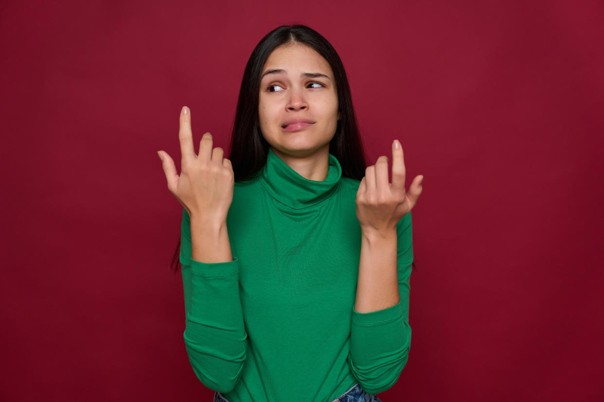 Woman in green turtleneck against maroon background holds up hands and has an expression of dread or confusion.