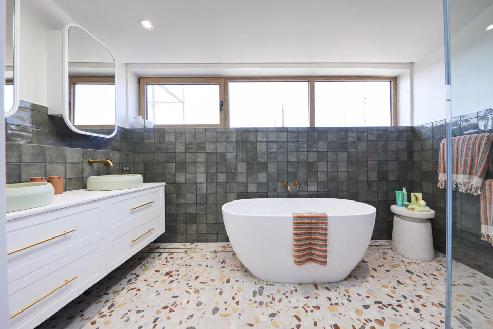 A wide shot of the bathroom with a vanity on the left, a bath on the far wall and textured tiles 