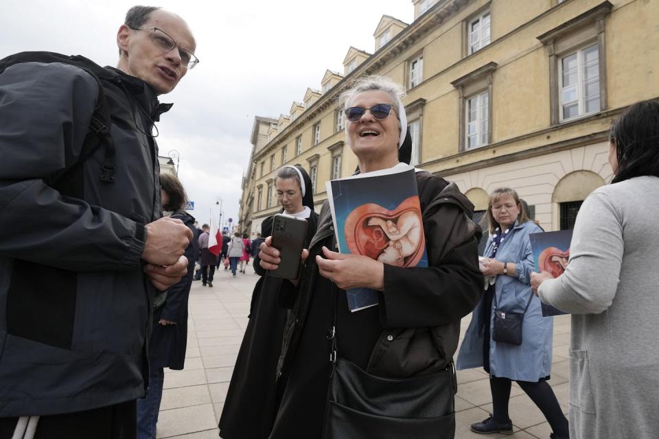 A woman in sunglasses and a nun's clothing holds an image of a child in utero as she speaks with a man on the street.
