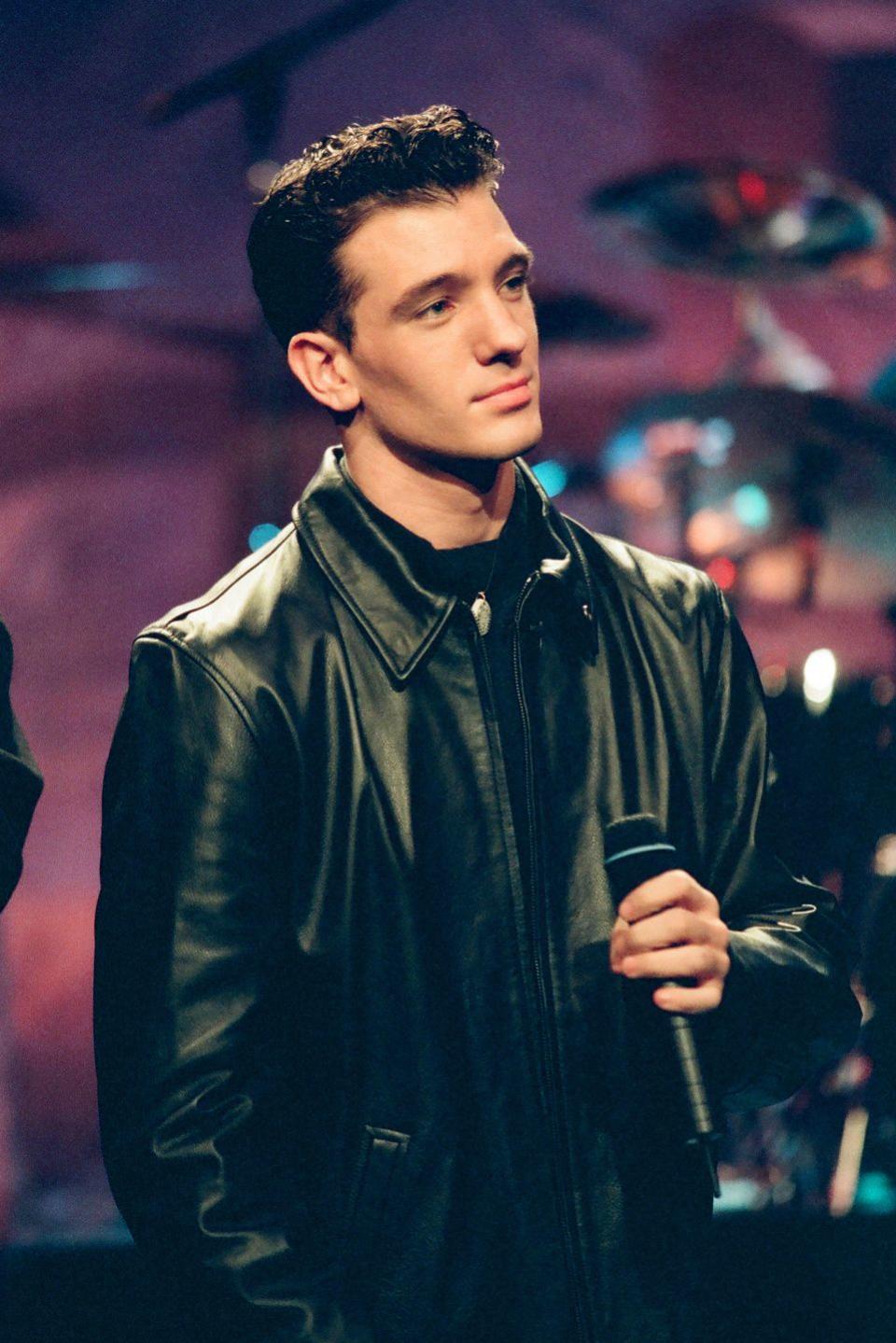 jc chasez holds a microphone at his chest and looks to the right, he wears a black jacket over a black shirt