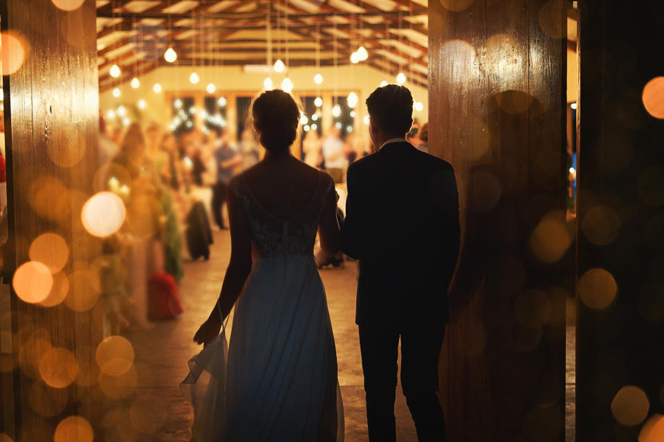 A couple stands arm in arm, silhouetted against a warmly lit venue filled with people and string lights, evoking a romantic ambiance