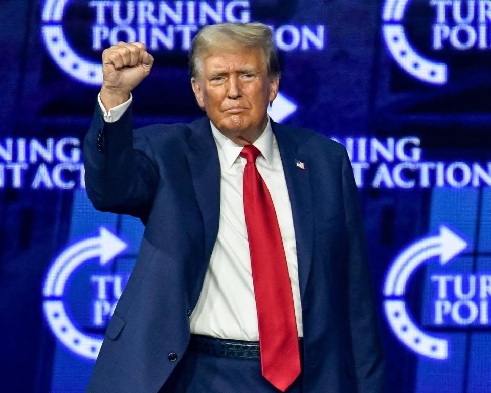 Donald Trump raises his fist while on stage at a Turning Point Action event, wearing a suit with a red tie