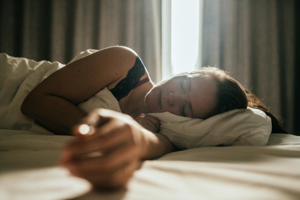 A woman is peacefully sleeping on a bed, resting her head on a pillow with her right arm outstretched. Sunlight shines through the curtains behind her