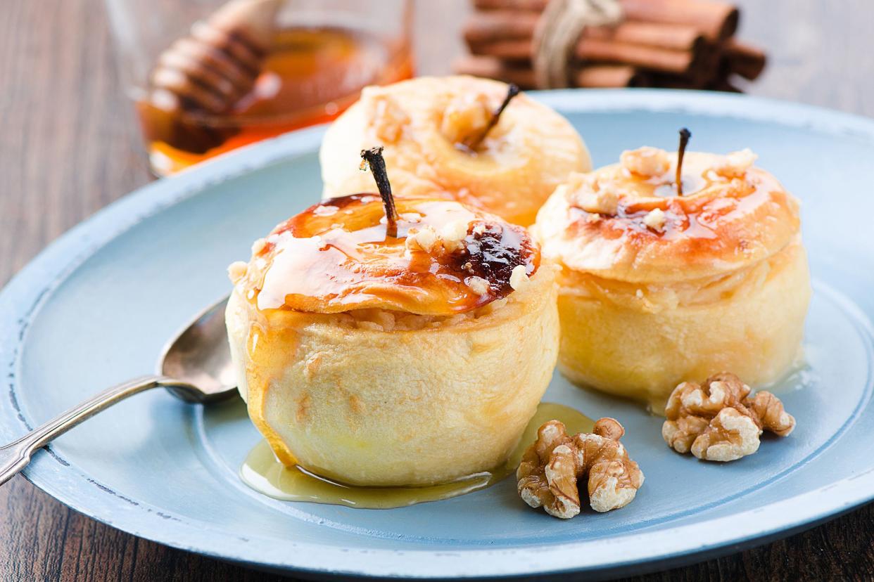 Three baked cinnamon stuffed apples on a light blue plate with a spoon with a glass jar of honey and a roll of cinnamon sticks in the background