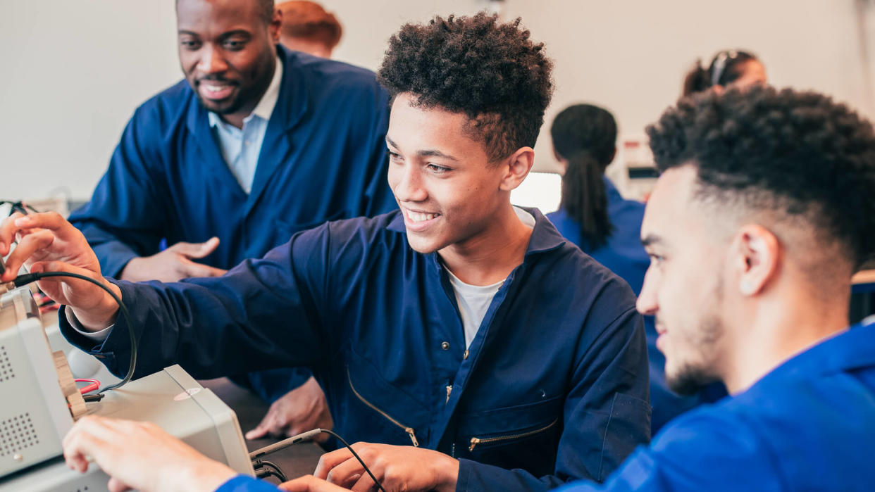 Two mixed race engineering students work on a group project together in a workshop whilst their tutor watches in the background.