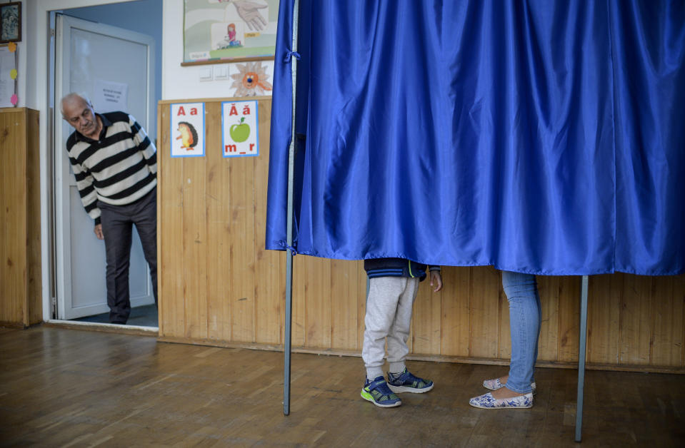 A man watches a mother casting her vote next to her child in a voting booth, in Ciorogarla, Romania, Sunday, Oct. 7, 2018. Romanians are voting Sunday around the country for a second day on a constitutional amendment that would make it harder to legalize same-sex marriage. (AP Photo/Andreea Alexandru)