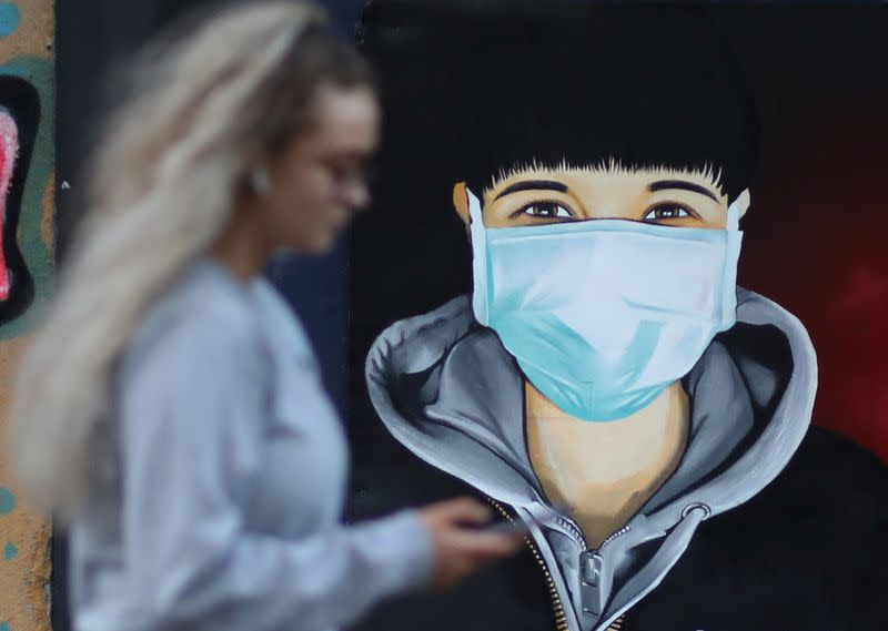 A woman checks her mobile phone as she walks past a mural depicting a man wearing a face mask as the spread of the coronavirus disease (COVID-19) continues, in Sale