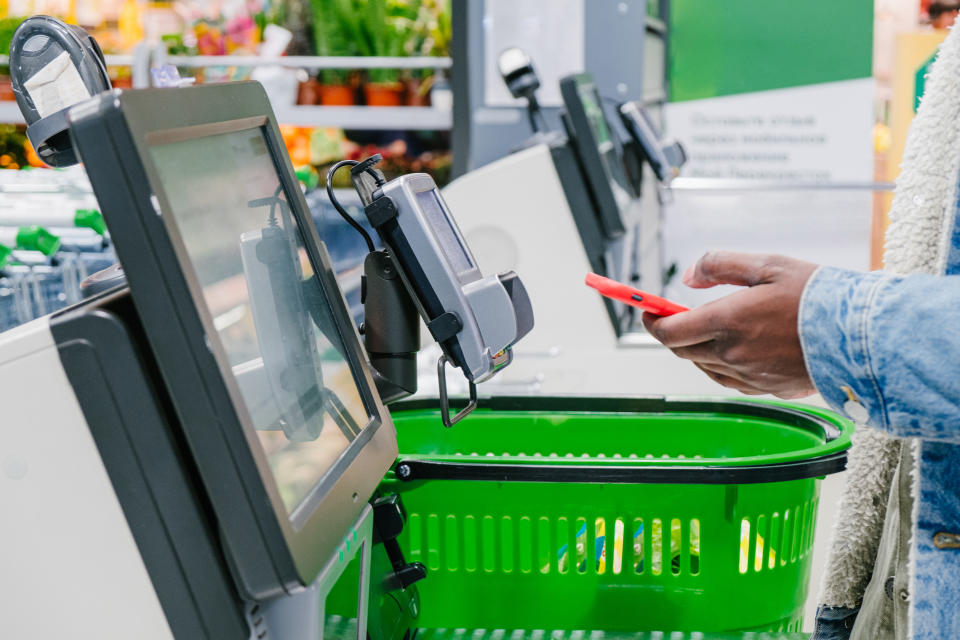 Person wearing warm denim jacket uses smartphone to pay for purchase at self-checkout point in grocery store.