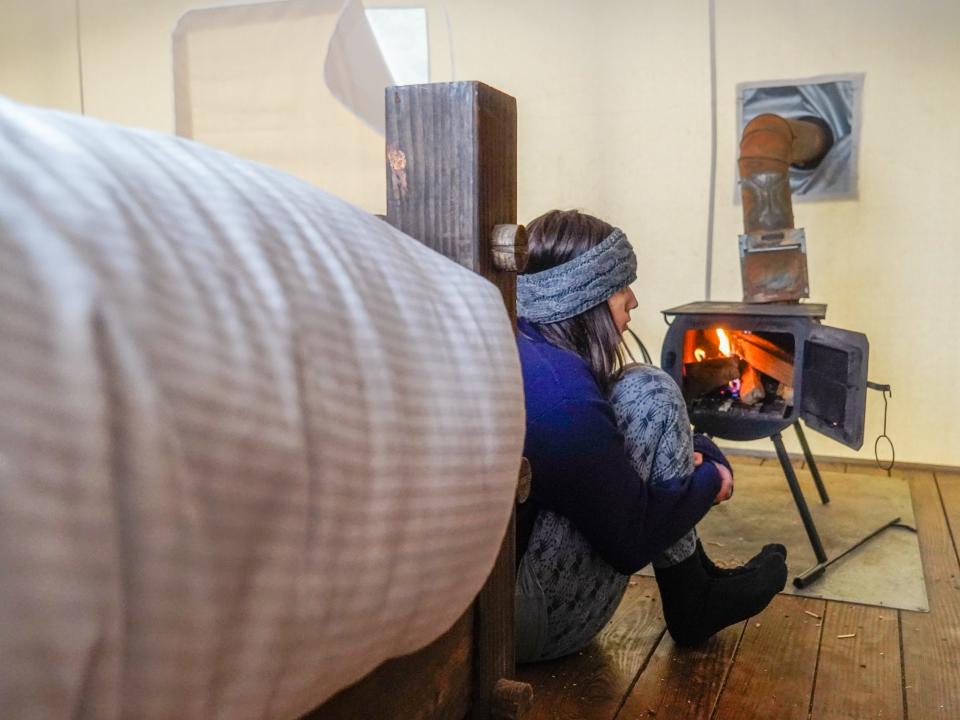 The author sits with her knees up in front of a fire with a bed on the left side of the frame