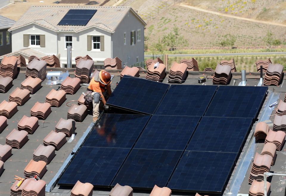 A worker places a solar panel on a roof surrounded by stacks of roof tiles.