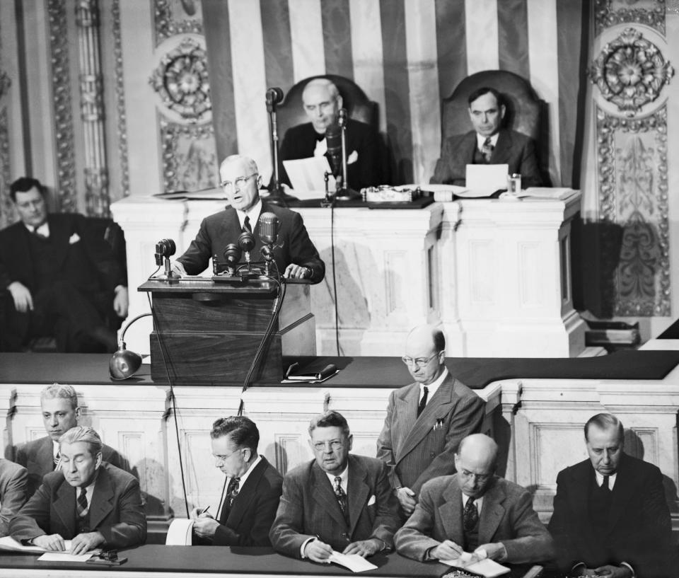 A historical political gathering features men in suits, including a speaker at a podium, others seated and writing in front and behind the speaker, and an American flag backdrop