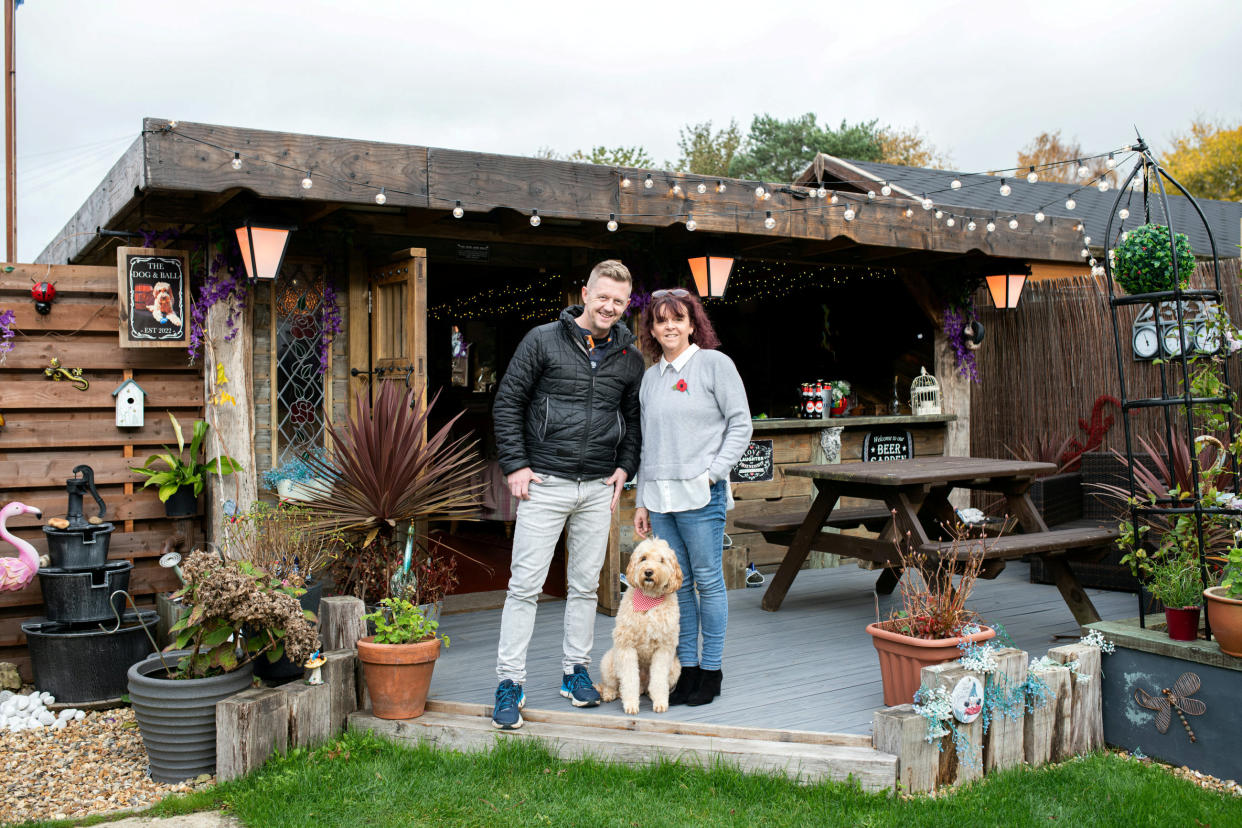 John & Anita Simmons with dog Bertie at their pub shed called The Dog & Ball in Horndean, Hampshire.  An incredible back garden boozer made entirely from recycled materials has been crowned Britain's best Pub Shed of the Year.  See SWNS story SWMRshed.  John Simmons, spent more than a year constructing the amazing DIY man-cave in his garden in Portsmouth, Hamps.   It features its own dart board, wooden dÃ©cor, countryside-pub style seating, a roof covered in fairy lights and even its own outdoor beer garden and decking area.   He built the miniature pub- called The Dog & Ball - from as much reclaimed material he could find from salvage yards and a Facebook page for pub shed enthusiasts.   John has now beat off more than a thousand entries to be named the owner of Britain's best Pub Shed 2022. 