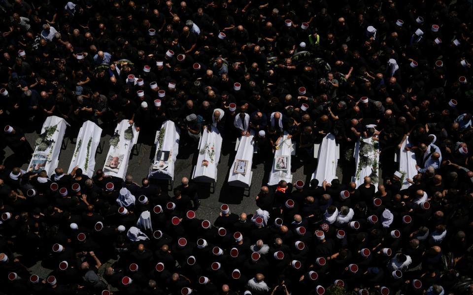 A row of ten white coffins is seen from high above, surrounded by men, many wearing traditional Druze hats