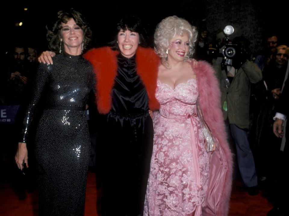 Jane Fonda in a black shiny gown, Lilly Tomlin in a black dress and red fur shawl, and Dolly Parton in a baby pink floral lace dress with a pink fur coat.