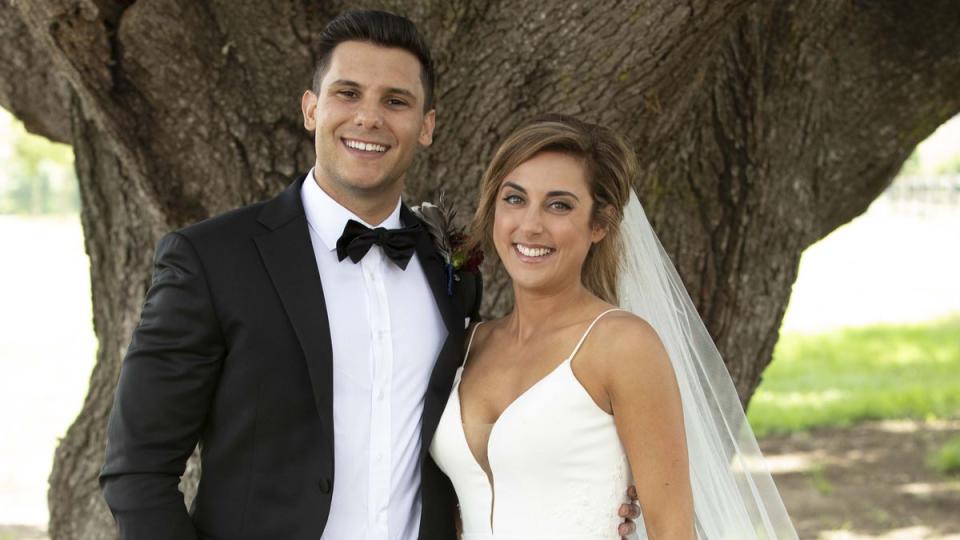 2021 MAFS couple Kerry and Johnny stand near a large tree on their show wedding day. He wears a dark suit and bow tie. She wears a strappy gown with plunging neckline and veil. Her streaked hair is tied back.