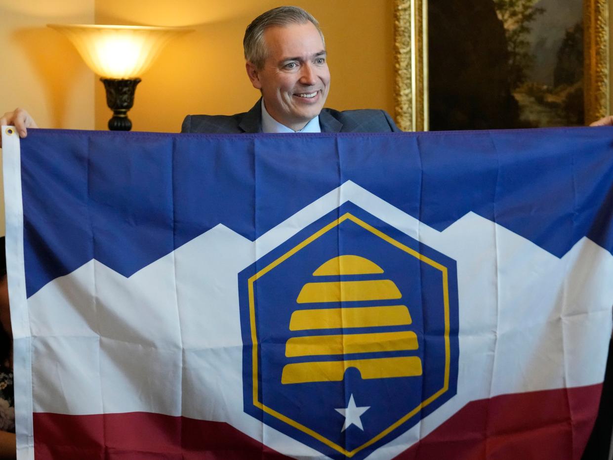 Sen. Daniel McCay holding up the state's new flag design, which features a beehive in front of striped blue, white, and red background with five mountain peaks.