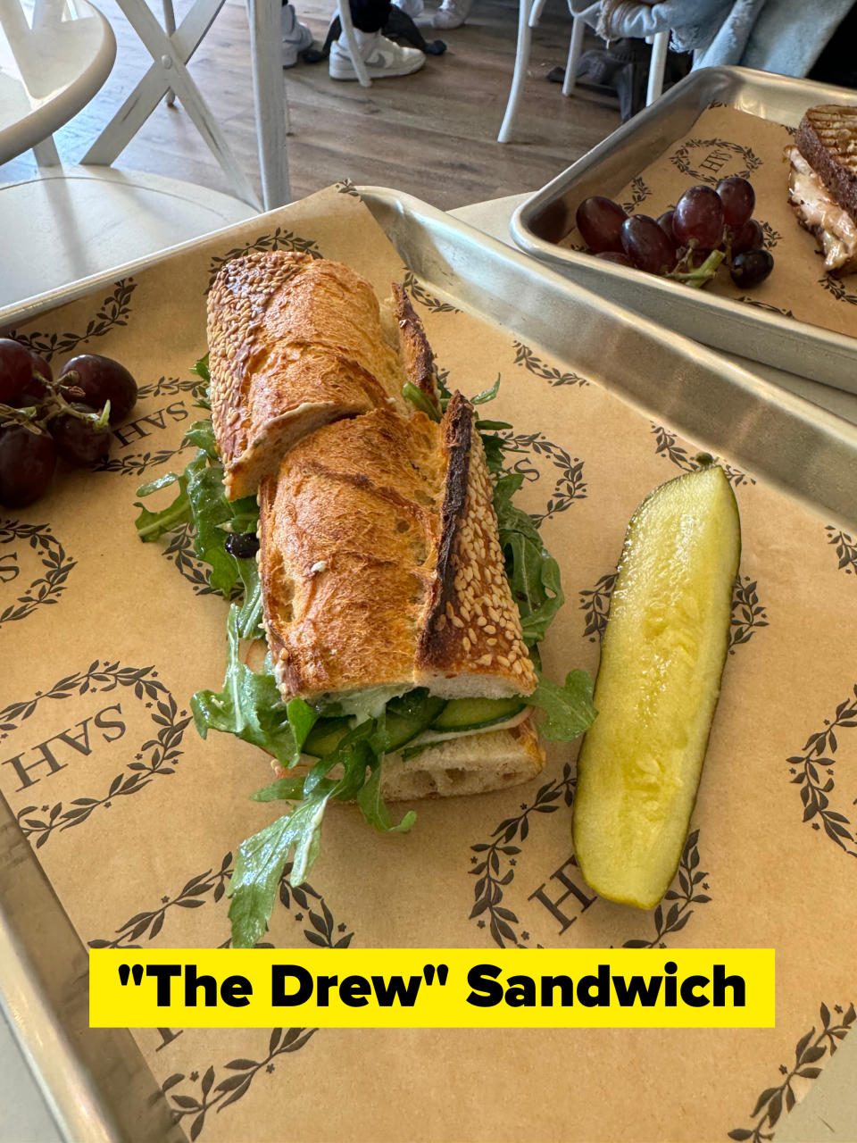 A sandwich on a tray with a pickle spear and a side of grapes at a restaurant
