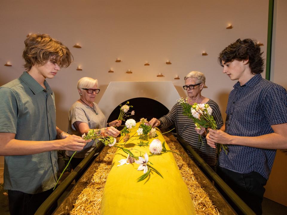 Two young men and two older women are placing flowers on a dummy body wrapped in a yellow shroud placed on wood chips, that is about to be loaded into a circular decomposition pod.