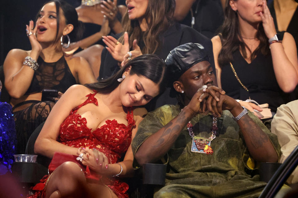 A smiling Selena resting her head on Rema's shoulder in the audience