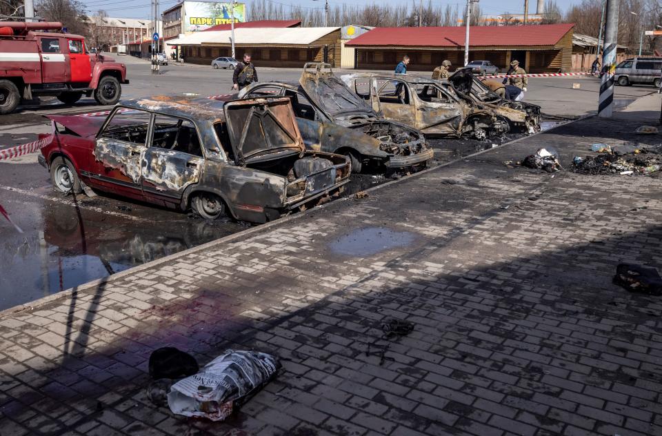 Four people stand near destroyed, calcinated cars in a parking area by a sidewalk.